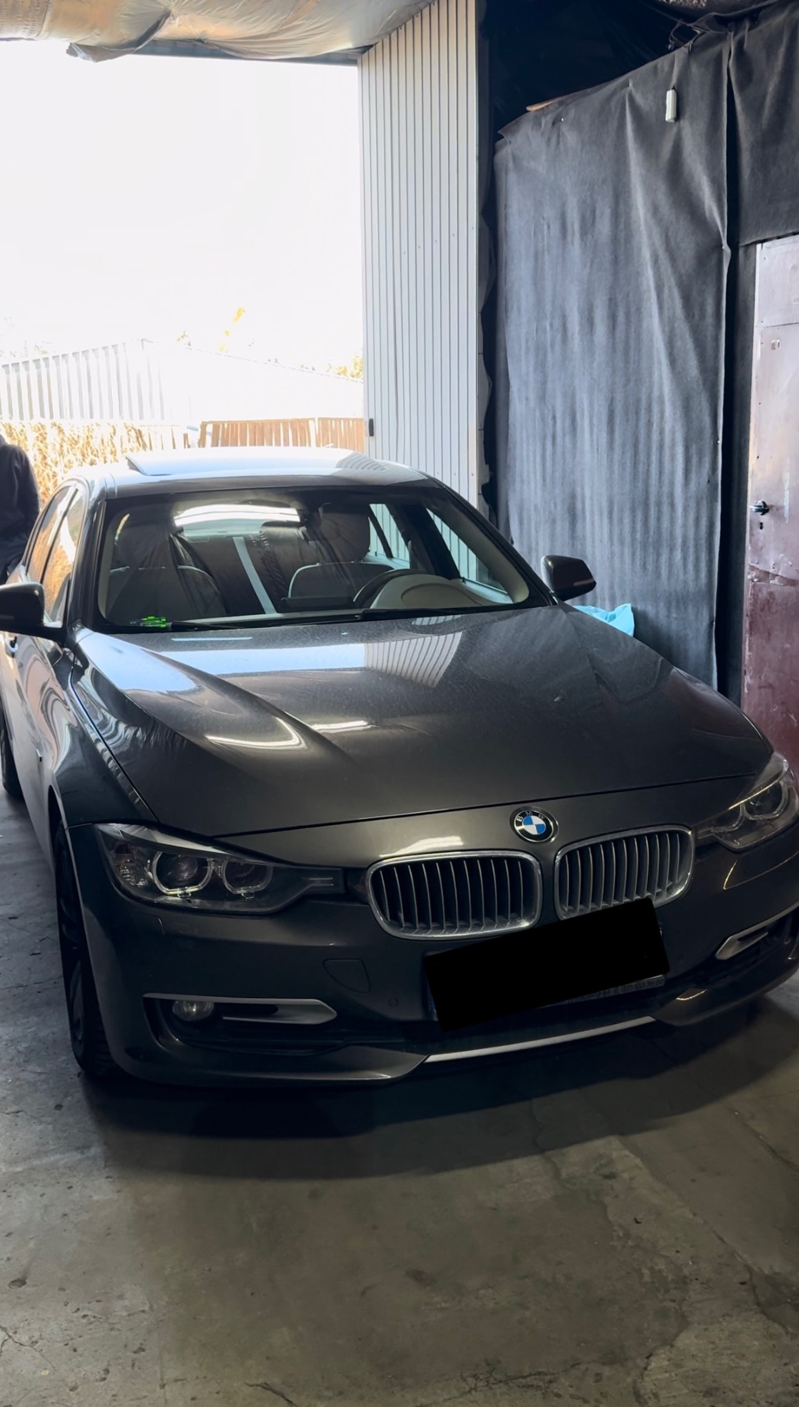 Szary samochód BMW serii 3 z otwartym szyberdachem, zaparkowany w warsztacie z szarą podłogą i metalowymi drzwiami garażowymi w tle.