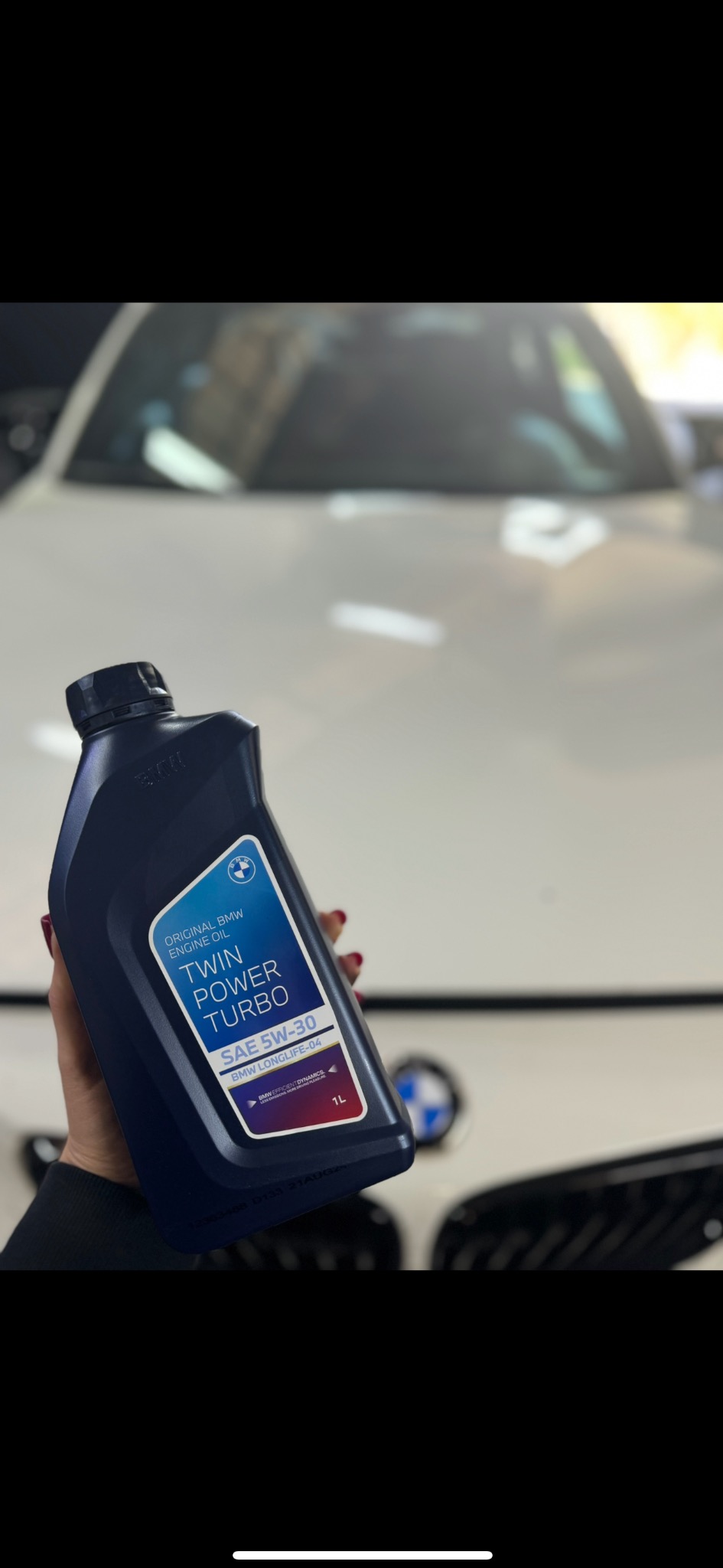 Oryginalny olej silnikowy BMW Twin Power Turbo SAE 5W-30 Longlife-04, trzymany w dłoni na tle białego samochodu BMW, widoczne logo BMW na opakowaniu i na samochodzie.