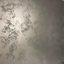 KAER Krzysztof Ratajczak - Ściana pokryta dekoracyjną, srebrzystą farbą z efektem chmur, subtelne przejścia tonalne, gładka powierzchnia z widoczną strukturą, oświetlenie podkreśla metaliczny połysk.