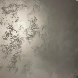 Ściana pokryta dekoracyjną, srebrzystą farbą z efektem chmur, subtelne przejścia tonalne, gładka powierzchnia z widoczną strukturą, oświetlenie podkreśla metaliczny połysk.