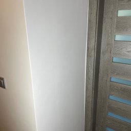 KAER Krzysztof Ratajczak - Świeżo pomalowana, gładka, biała ściana w rogu pomieszczenia, sąsiadująca z beżową ścianą z włącznikami oraz drewnianymi drzwiami z poziomymi, błękitnymi wstawkami.