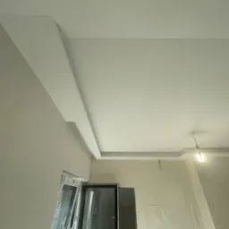 Wykończone gładzią ściany i sufit w jasnym wnętrzu z oknem. Widoczne listwy dekoracyjne pod sufitem i lampa. Ściany przygotowane do malowania.