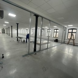 KAER Krzysztof Ratajczak - Montaż metalowej konstrukcji ścianki działowej w przestronnym, jasnym pomieszczeniu z kolumnami. Widoczne narzędzia i drabina. Etap prac przed obudową płytami.