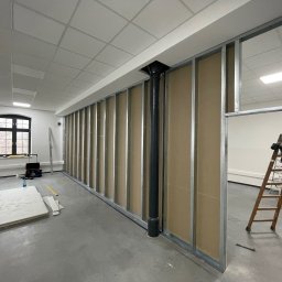 KAER Krzysztof Ratajczak - Montaż ścianki działowej z płyt gipsowo-kartonowych w biurze. Widoczne profile, płyty, drabina i narzędzia. Czarna rura pionowa wkomponowana w konstrukcję.