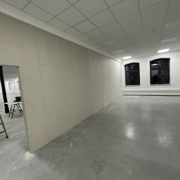 KAER Krzysztof Ratajczak - Wnętrze w trakcie prac: ściany z płyt kartonowo-gipsowych, widoczna drabina i narzędzia. Szare podłoże, dwa okna z czarną ramą.