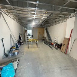 KAER Krzysztof Ratajczak - Montaż metalowej konstrukcji sufitu podwieszanego w surowym wnętrzu. Widoczne narzędzia i materiały budowlane. Prace wykończeniowe w trakcie realizacji.