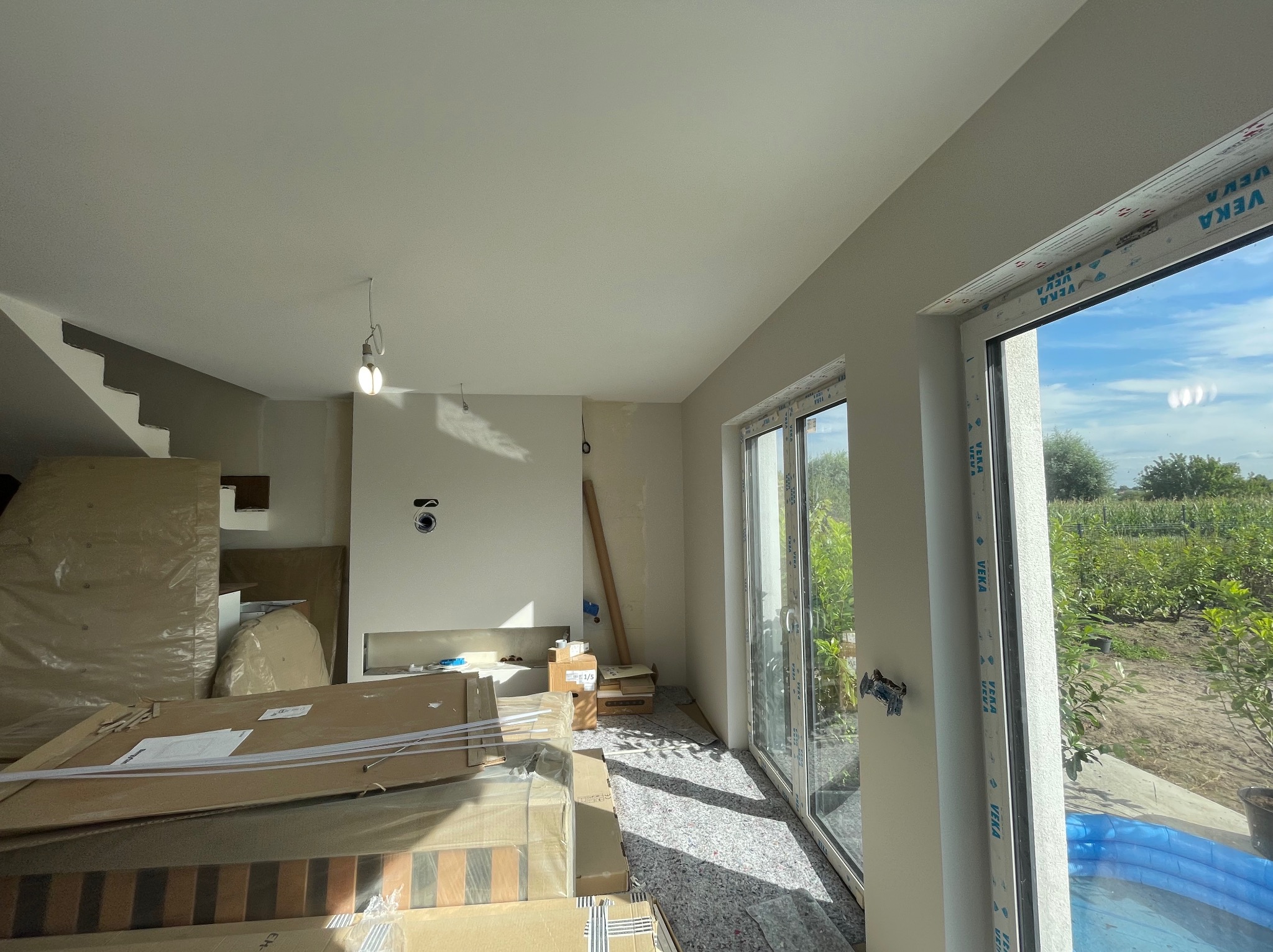 Jasne wnętrze w trakcie wykończenia: białe ściany, okna z widokiem na zieleń, podłoga zabezpieczona folią, kartony z materiałami budowlanymi i surowe schody.
