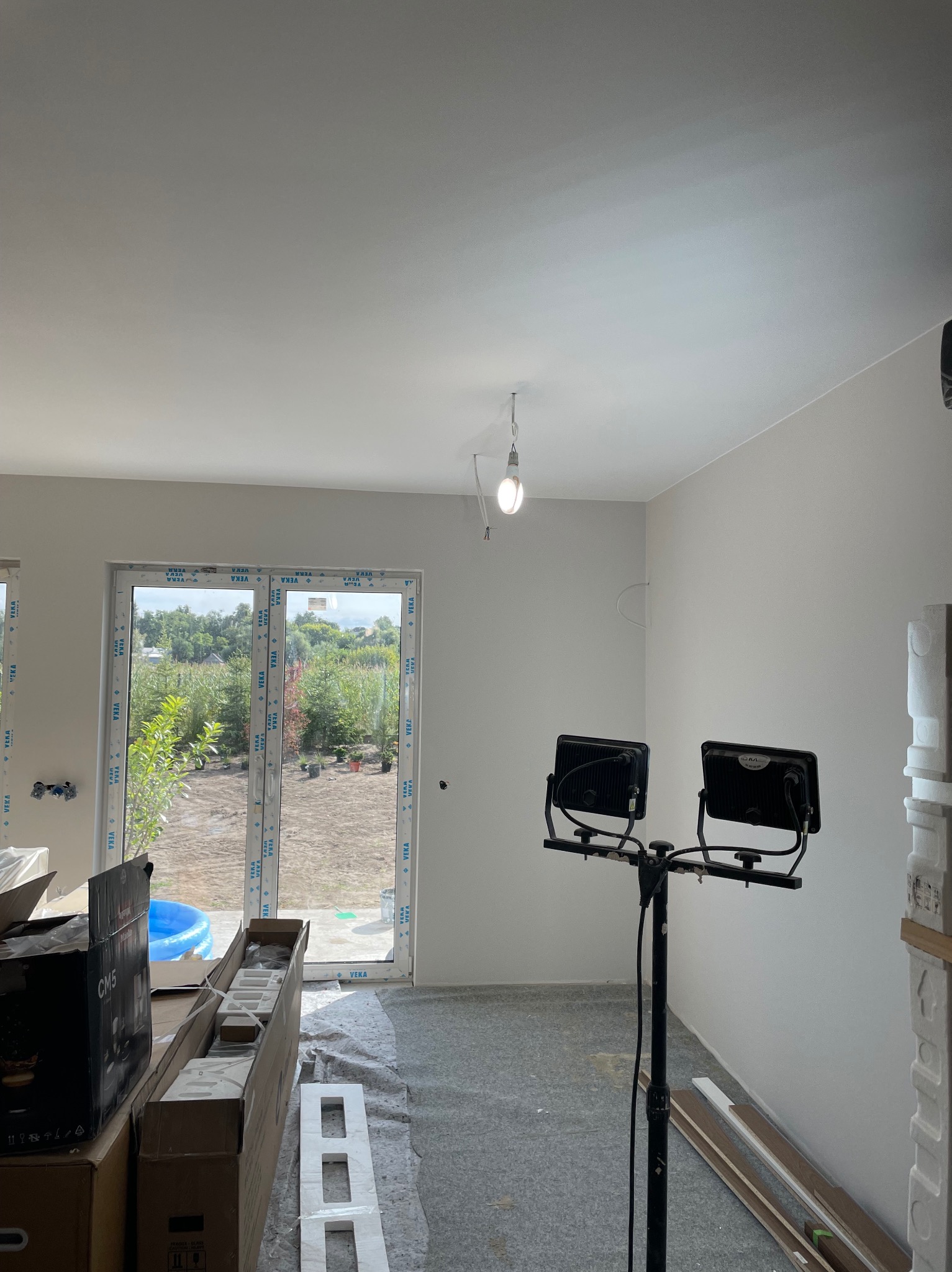 Wnętrze w trakcie prac wykończeniowych: gładkie, białe ściany, podłoga zabezpieczona folią, lampa budowlana na statywie, okno z widokiem na zieleń.