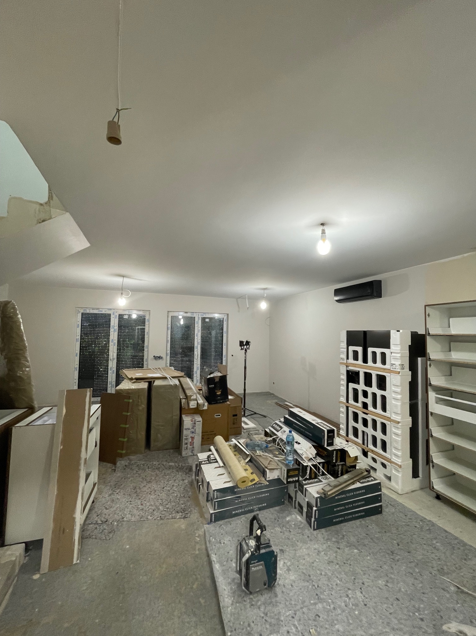 Wnętrze w trakcie wykończenia: puste ściany, okna z roletami, kartony, narzędzia i materiały podłogowe. Widoczny surowy stan i przygotowanie do prac wykończeniowych.