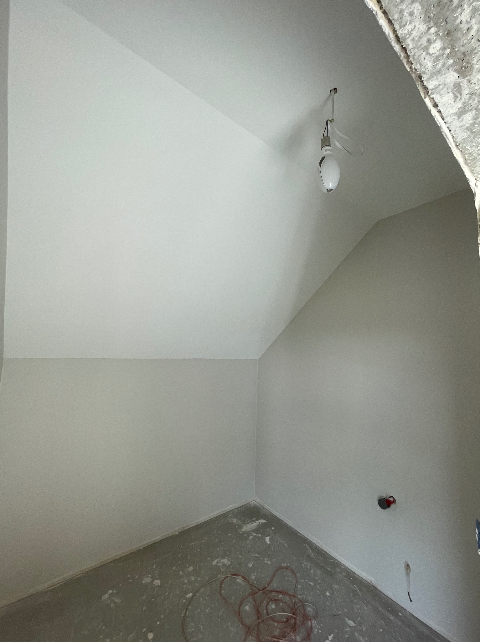 Wnętrze poddasza po szpachlowaniu ścian na gładko, przygotowane do malowania. Widoczne gniazdko elektryczne i kabel na podłodze. Surowy fragment ściany przy suficie.