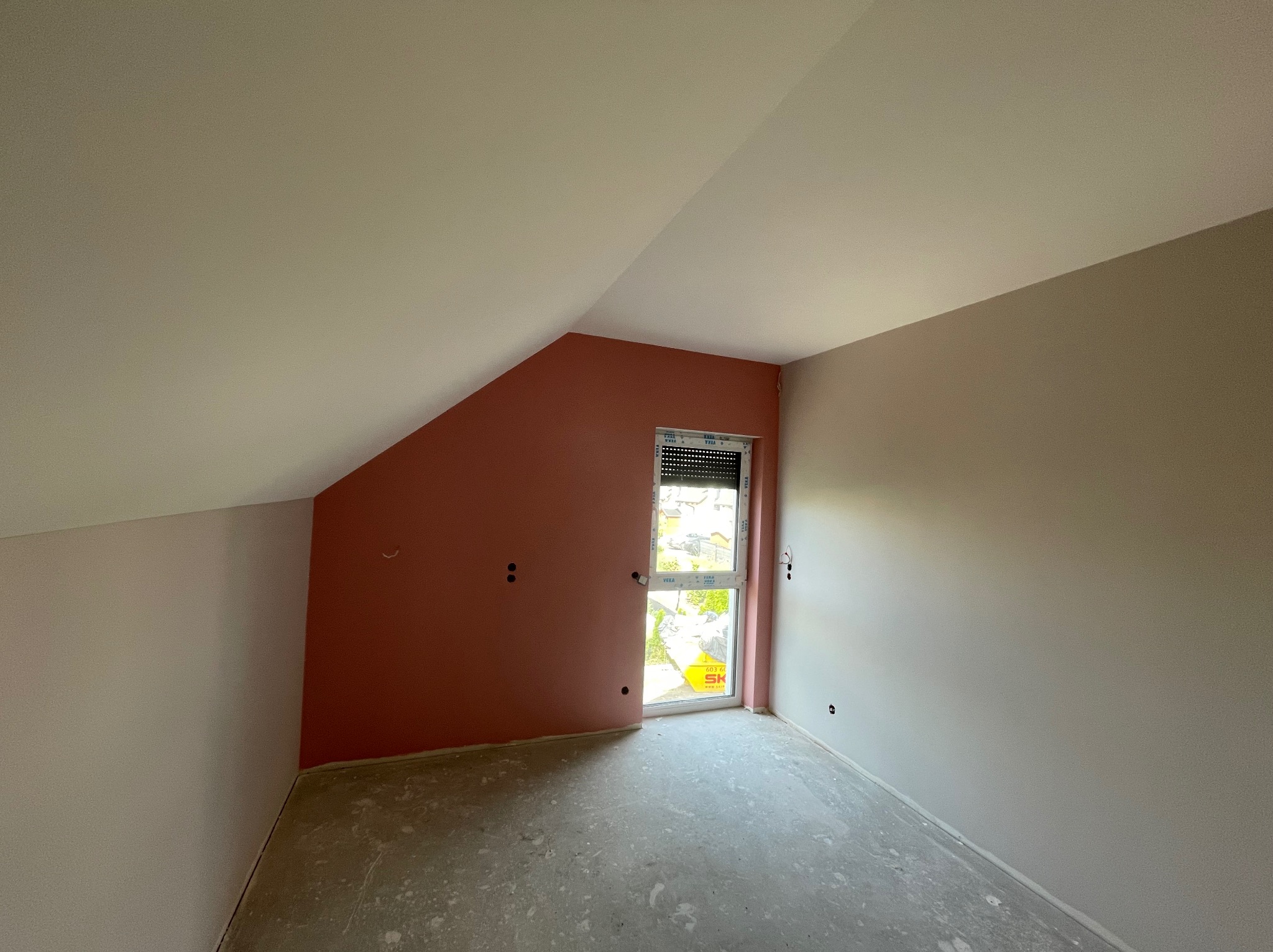 Pokój na poddaszu po szpachlowaniu, z oknem i ścianami w odcieniach beżu i różu. Podłoga surowa, przygotowana do dalszych prac wykończeniowych.