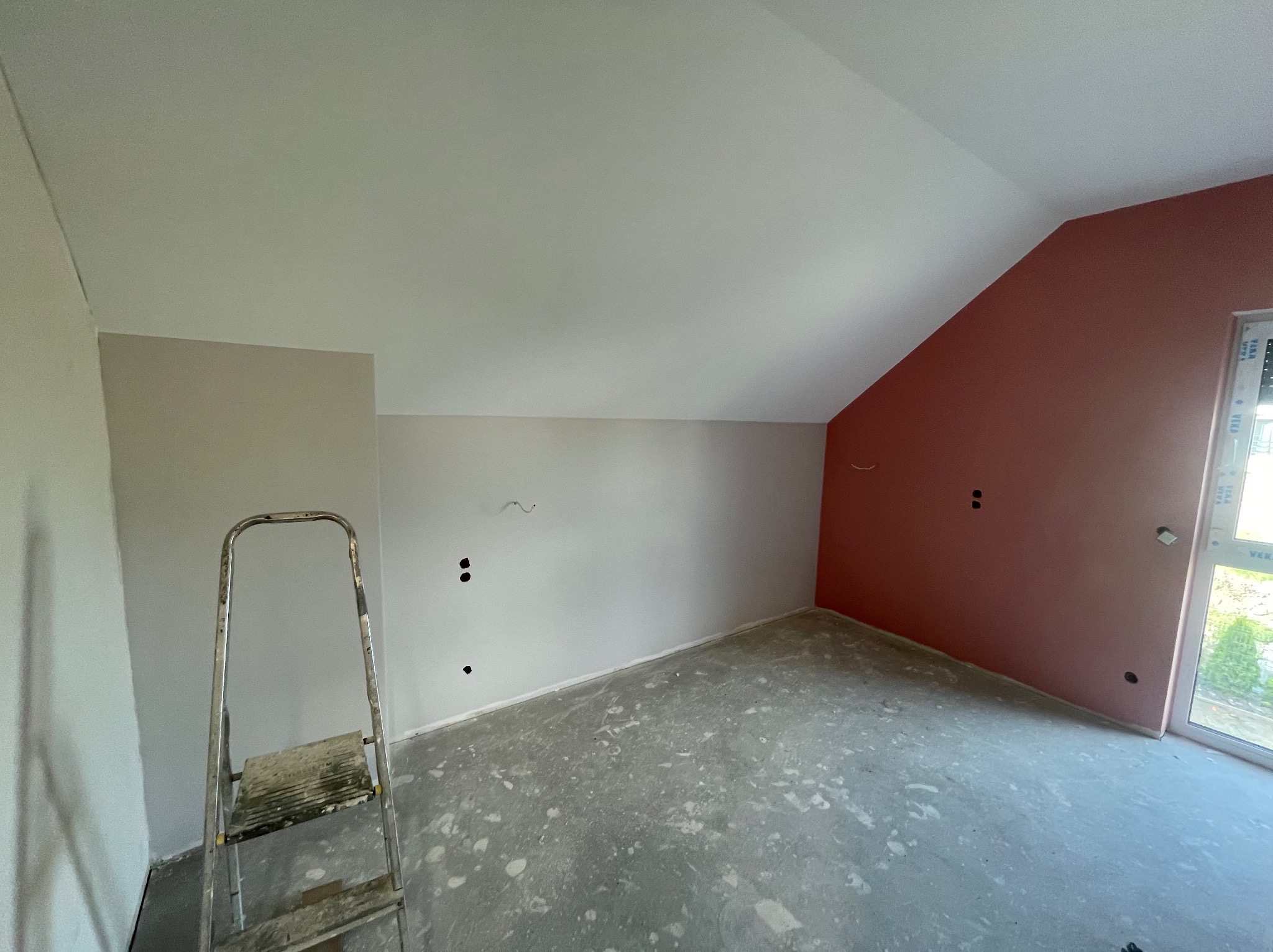 Pokój po szpachlowaniu, ściany częściowo pomalowane na różowo, widoczna drabina malarska i surowa podłoga. Przygotowania do wykończenia poddasza.