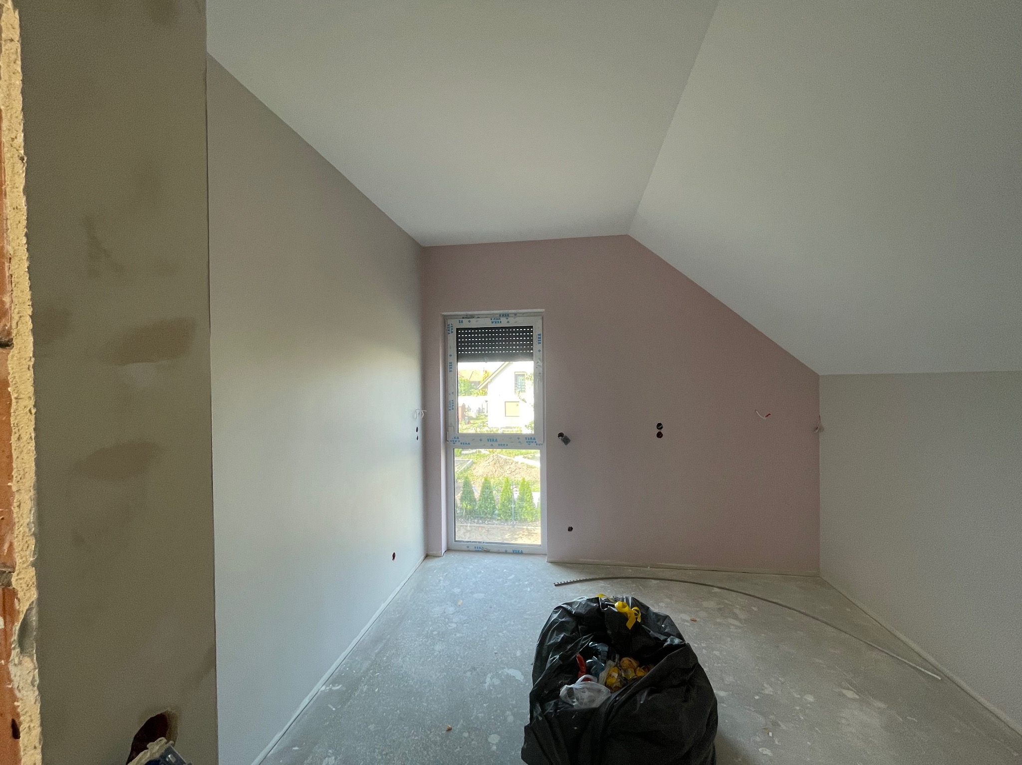 Pokój na poddaszu po szpachlowaniu ścian, z oknem i workiem na śmieci. Ściany w odcieniach szarości i różu, sufit biały. Widoczne ślady po gładzi na podłodze.