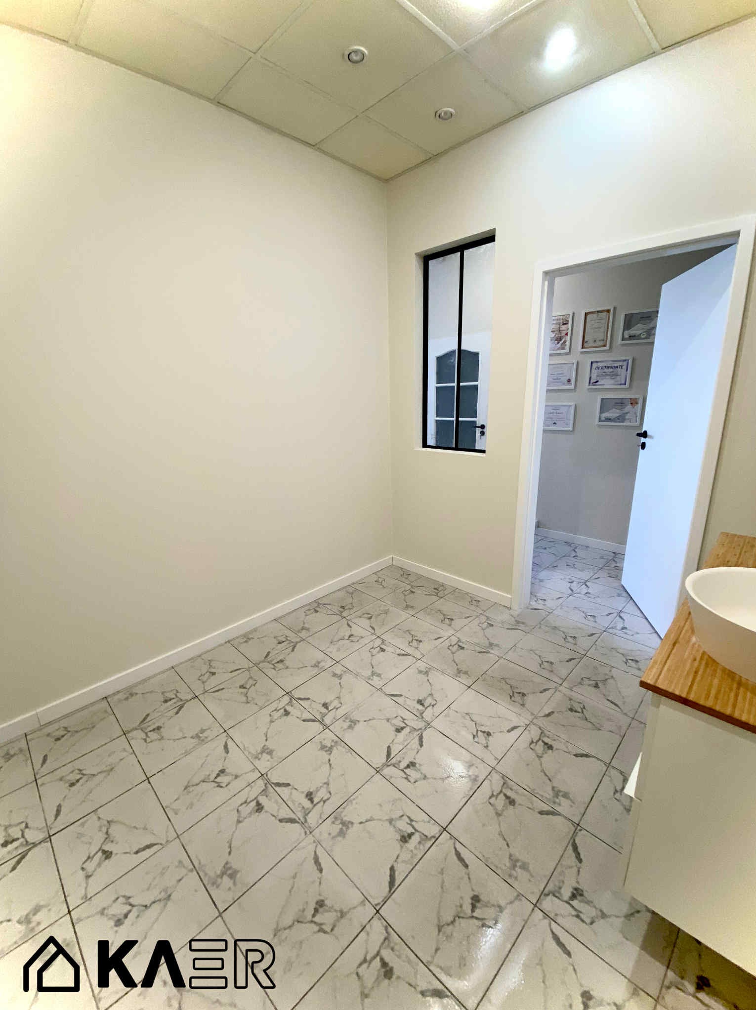 Wnętrze po remoncie: jasne ściany, marmurowe płytki podłogowe, okno z czarną ramą i widok na sąsiednie pomieszczenie z certyfikatami na ścianie.