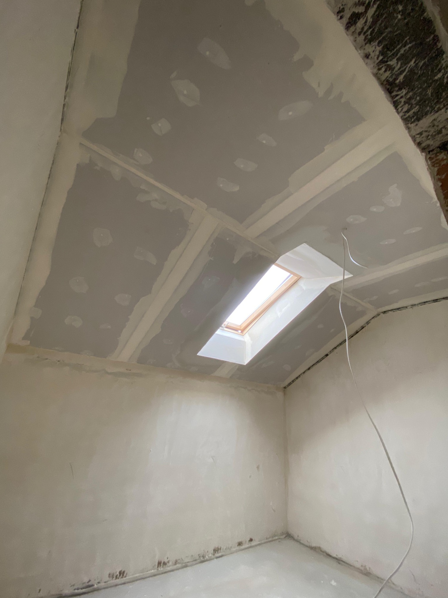 Pokój ze skośnym sufitem po montażu płyt gipsowo-kartonowych i szpachlowaniu, widoczne okno dachowe i wystające przewody. Ściany przygotowane do malowania.