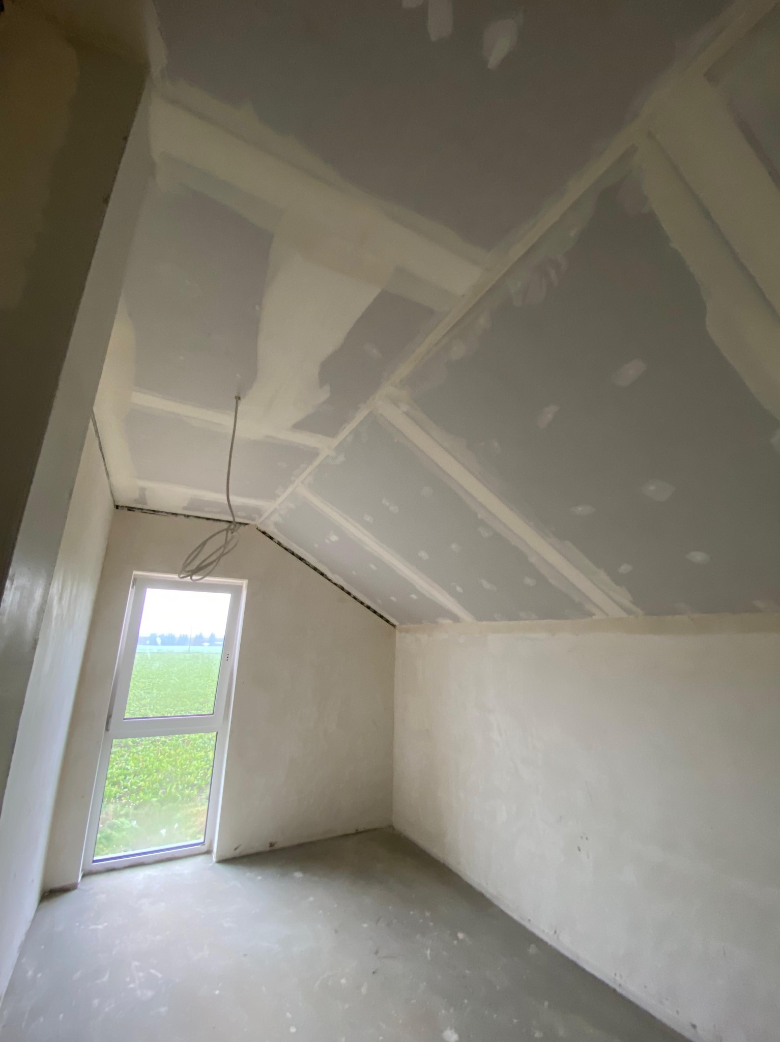 Pokój na poddaszu w trakcie prac wykończeniowych: ściany i skośny sufit pokryte płytami, widoczne łączenia i szpachlowanie. Okno z widokiem na zieleń.