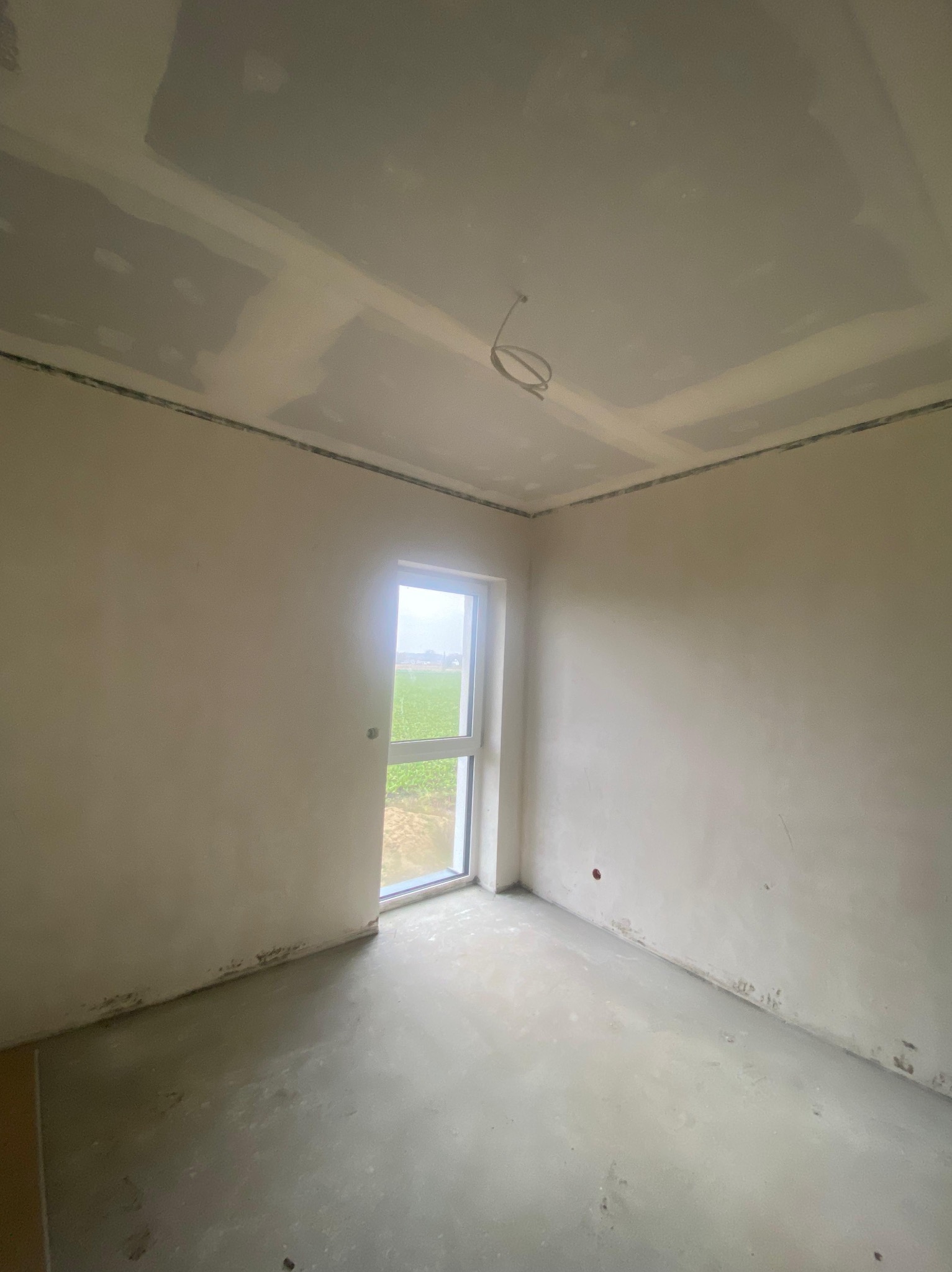 Pokój w trakcie wykończenia: ściany po szpachlowaniu, sufit z płyt karton-gips, widoczne przewody elektryczne. Okno z widokiem na zieleń.