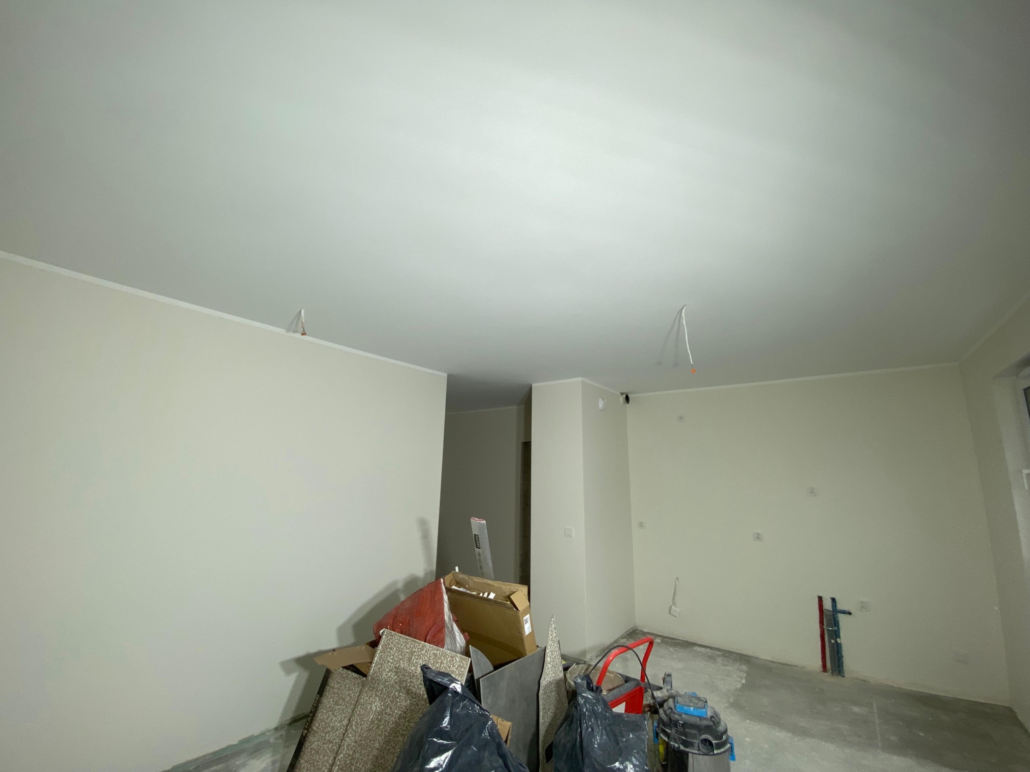 Wnętrze w trakcie prac wykończeniowych: świeżo pomalowane ściany w jasnym odcieniu, widoczne instalacje elektryczne i hydrauliczne, gruz i narzędzia na podłodze.