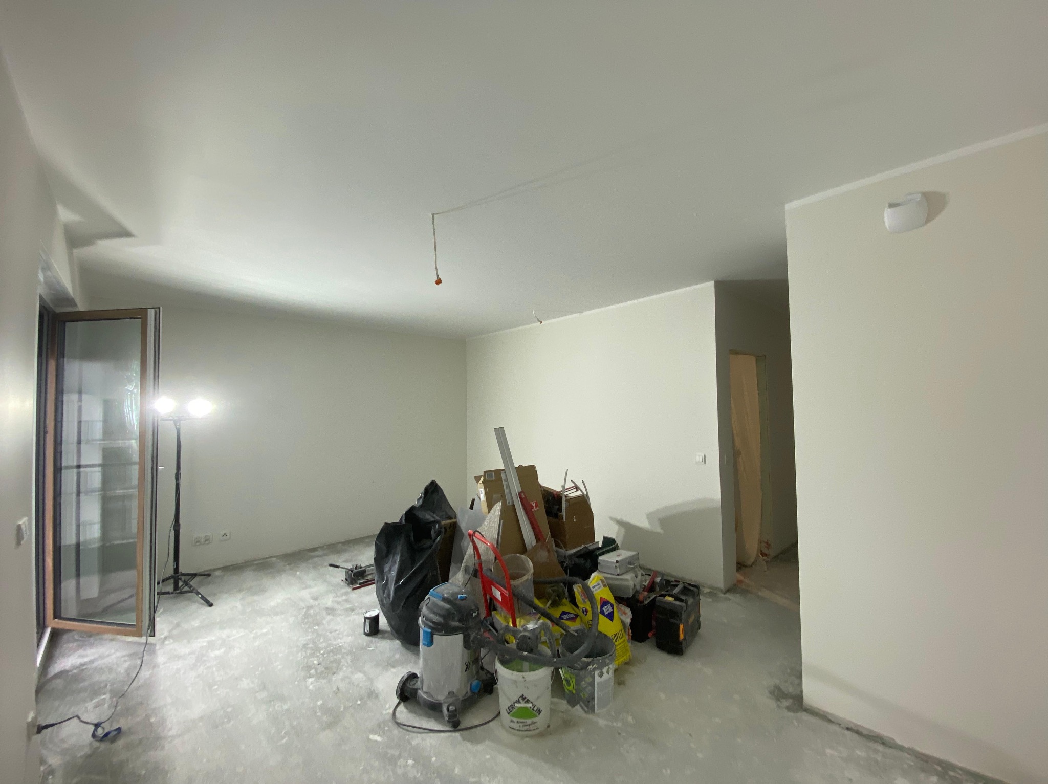 Wnętrze w trakcie prac wykończeniowych: widoczne narzędzia, worki i sprzęt budowlany na surowej podłodze, ściany przygotowane do dalszych prac. Oświetlenie halogenowe.