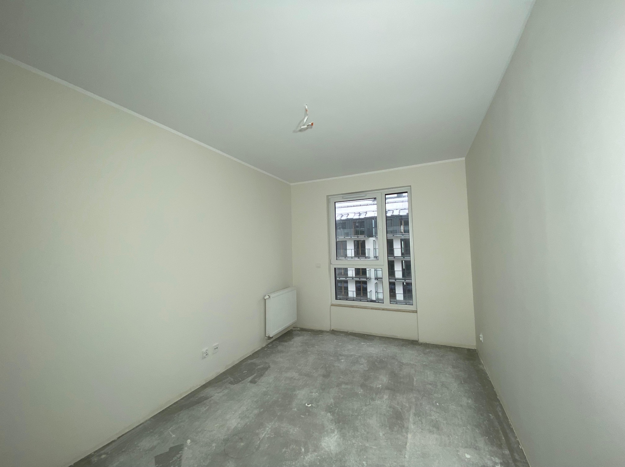 Jasne wnętrze pokoju w trakcie prac wykończeniowych. Widoczne ściany po szpachlowaniu, surowa podłoga i okno z widokiem na budynek.