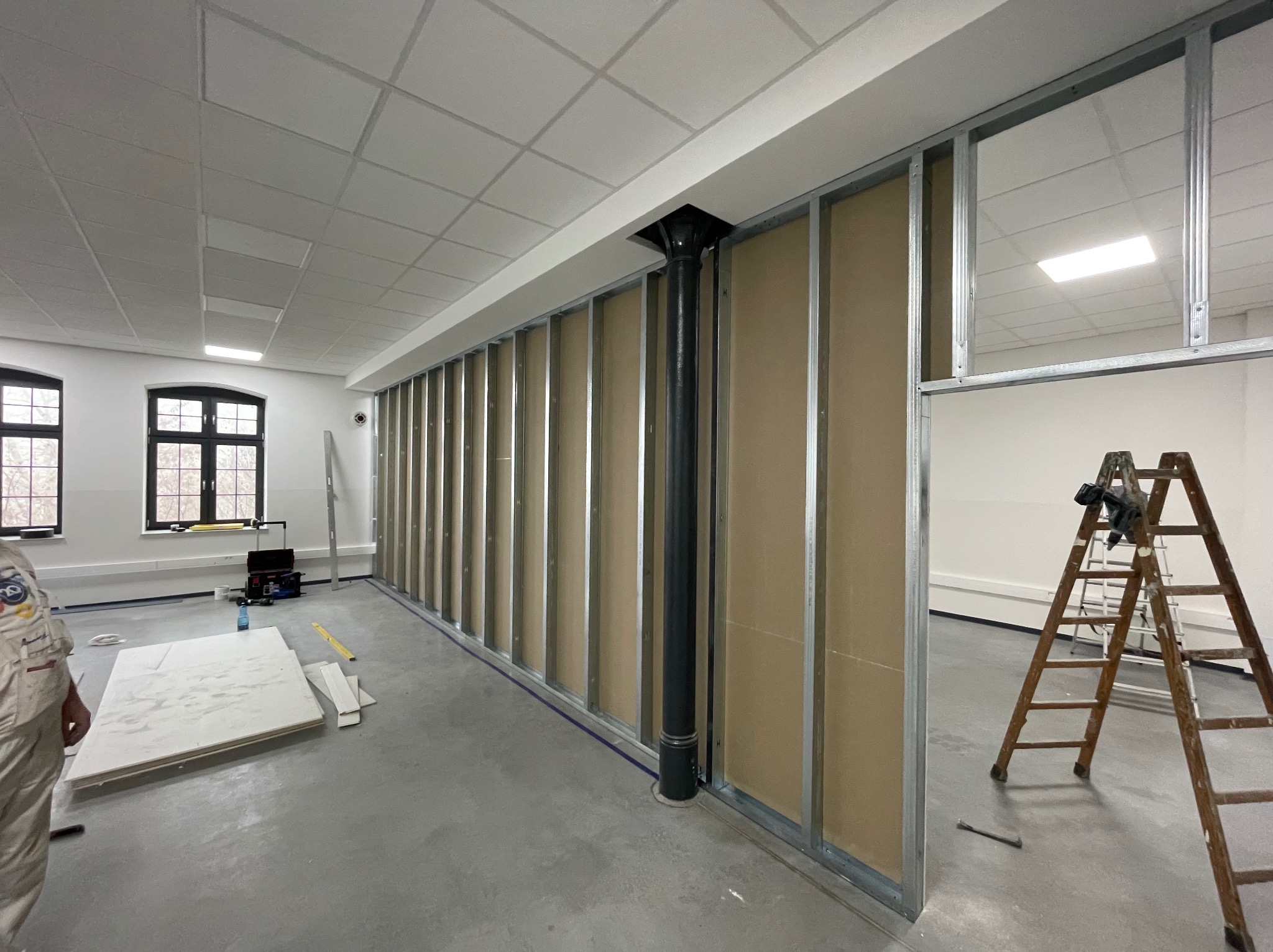 Montaż ścianki działowej z płyt gipsowo-kartonowych w biurze. Widoczne profile, płyty, drabina i narzędzia. Czarna rura pionowa wkomponowana w konstrukcję.