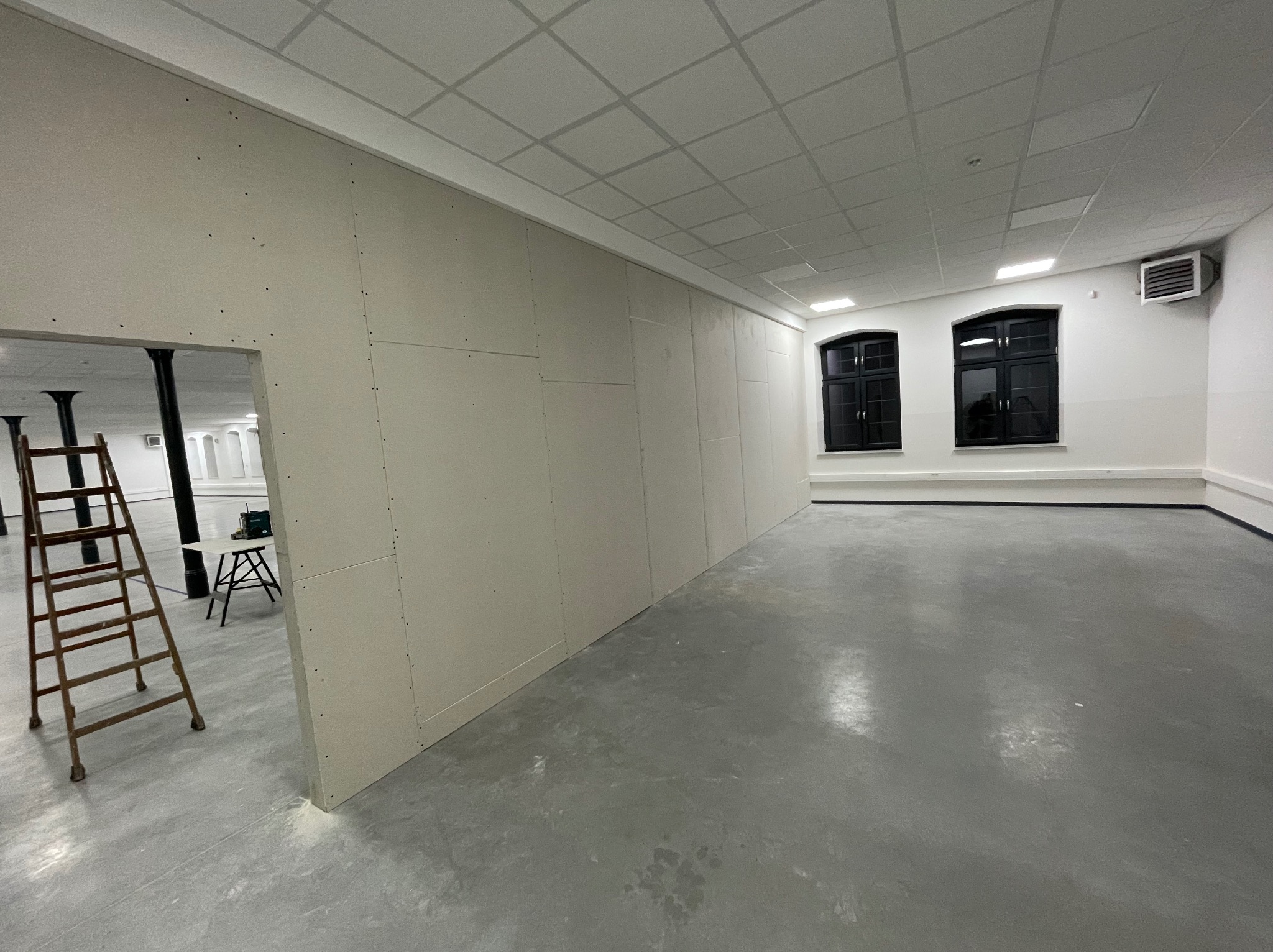 Wnętrze w trakcie prac: ściany z płyt kartonowo-gipsowych, widoczna drabina i narzędzia. Szare podłoże, dwa okna z czarną ramą.