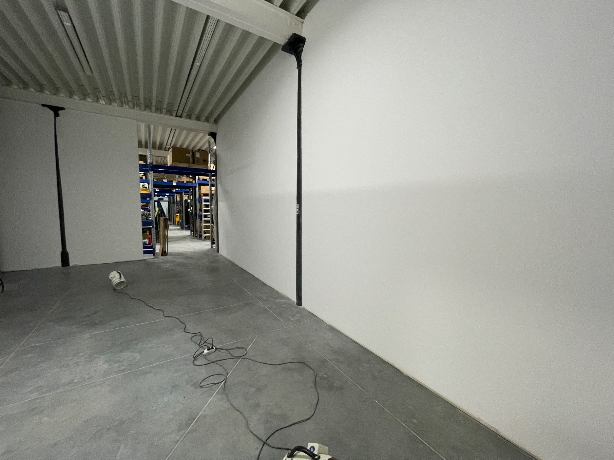 Biała, gładka ściana w magazynie po remoncie, widoczne szpachlowanie i gładzie. Szare płytki na podłodze, metalowa konstrukcja sufitu i regały w tle.