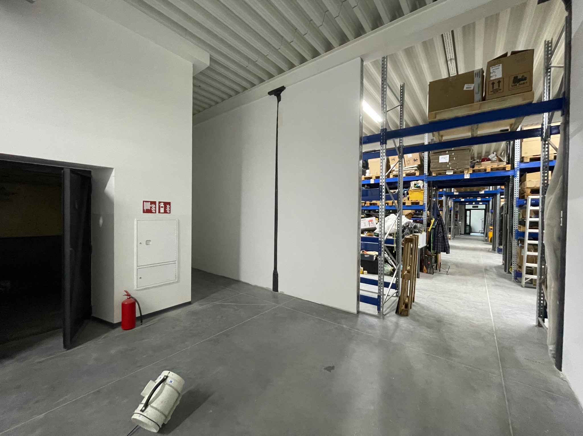 Wnętrze magazynu po remoncie: białe ściany, szara posadzka, regały magazynowe w tle. Widoczny sprzęt gaśniczy i narzędzie do malowania sufitów.