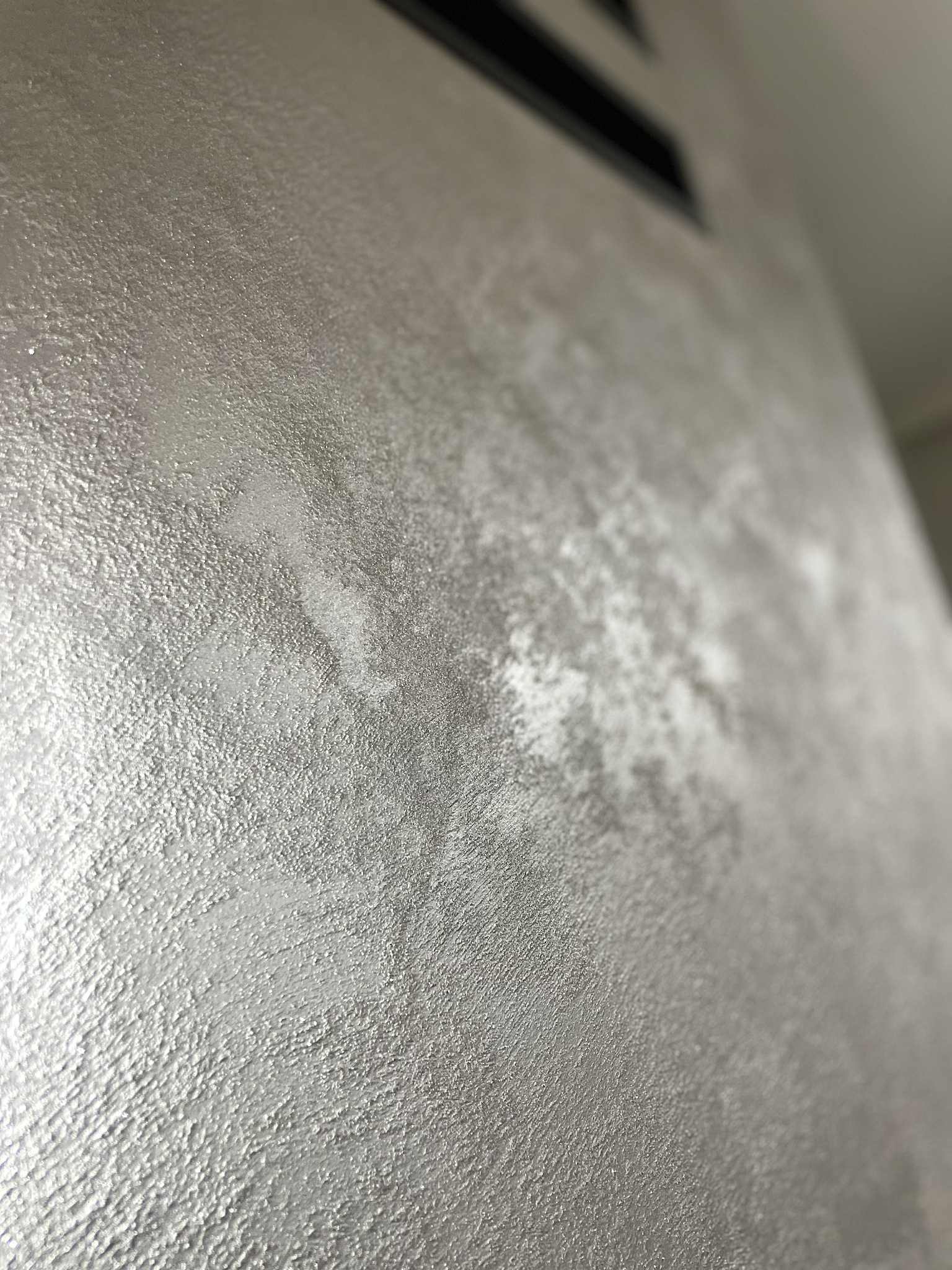 Szara, metaliczna ściana z wyraźną fakturą, oświetlona z góry, ukazana pod kątem. Widoczny fragment sufitu z czarnym elementem wentylacyjnym.