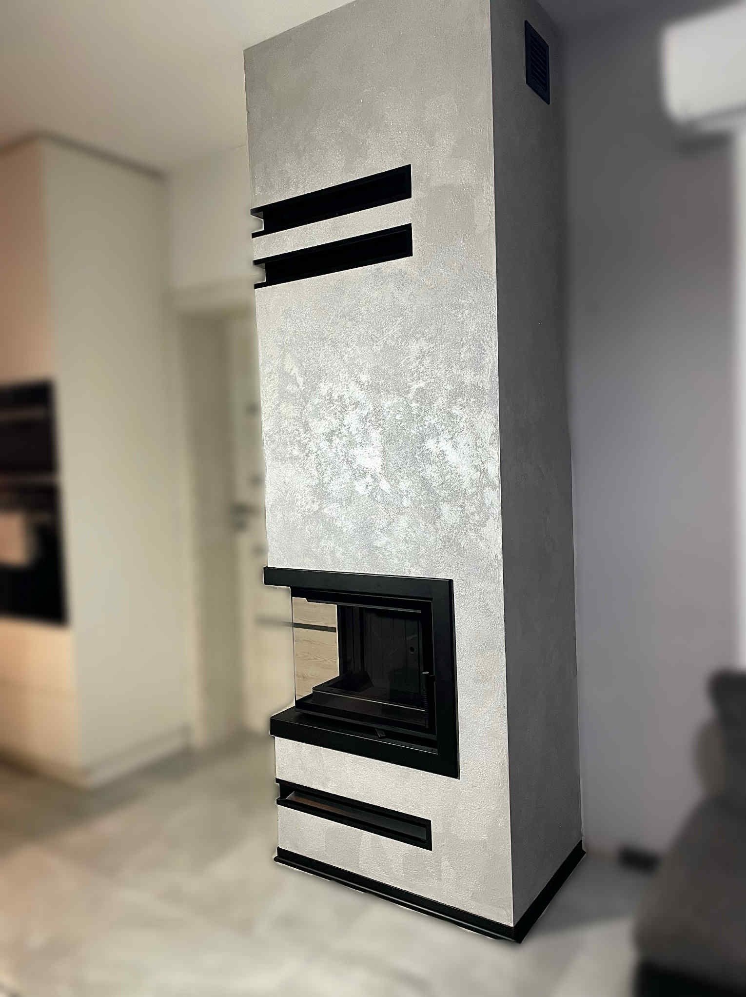 Nowoczesny kominek obudowany szarą, błyszczącą strukturą dekoracyjną z czarnymi, poziomymi wstawkami, w jasnym wnętrzu. Wykończenie w stylu minimalistycznym.