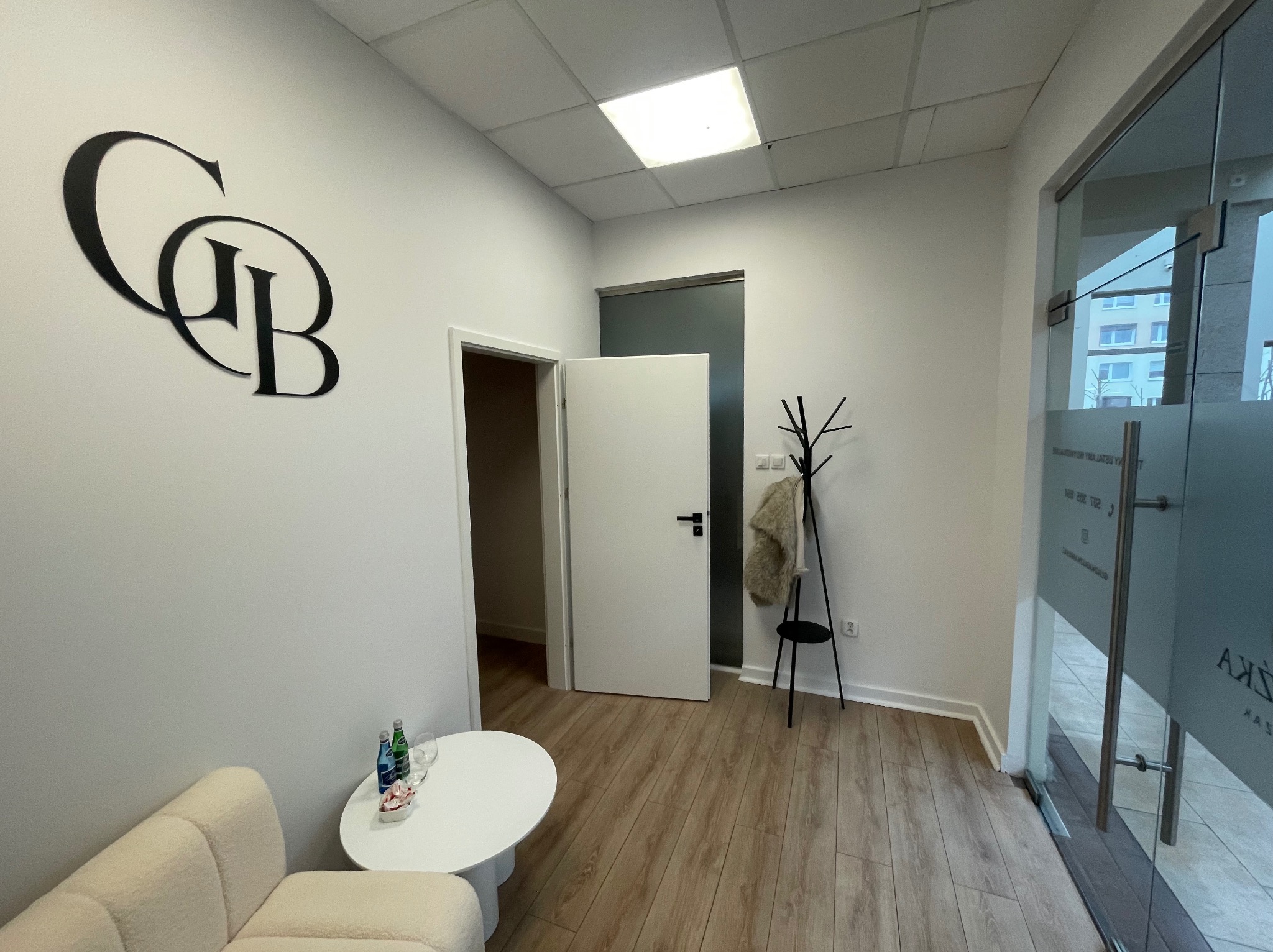 Jasne wnętrze biura z drewnianą podłogą, białymi ścianami i nowoczesnym wystrojem. Widoczny fragment sofy, stolik, wieszak na ubrania i logo firmy na ścianie.
