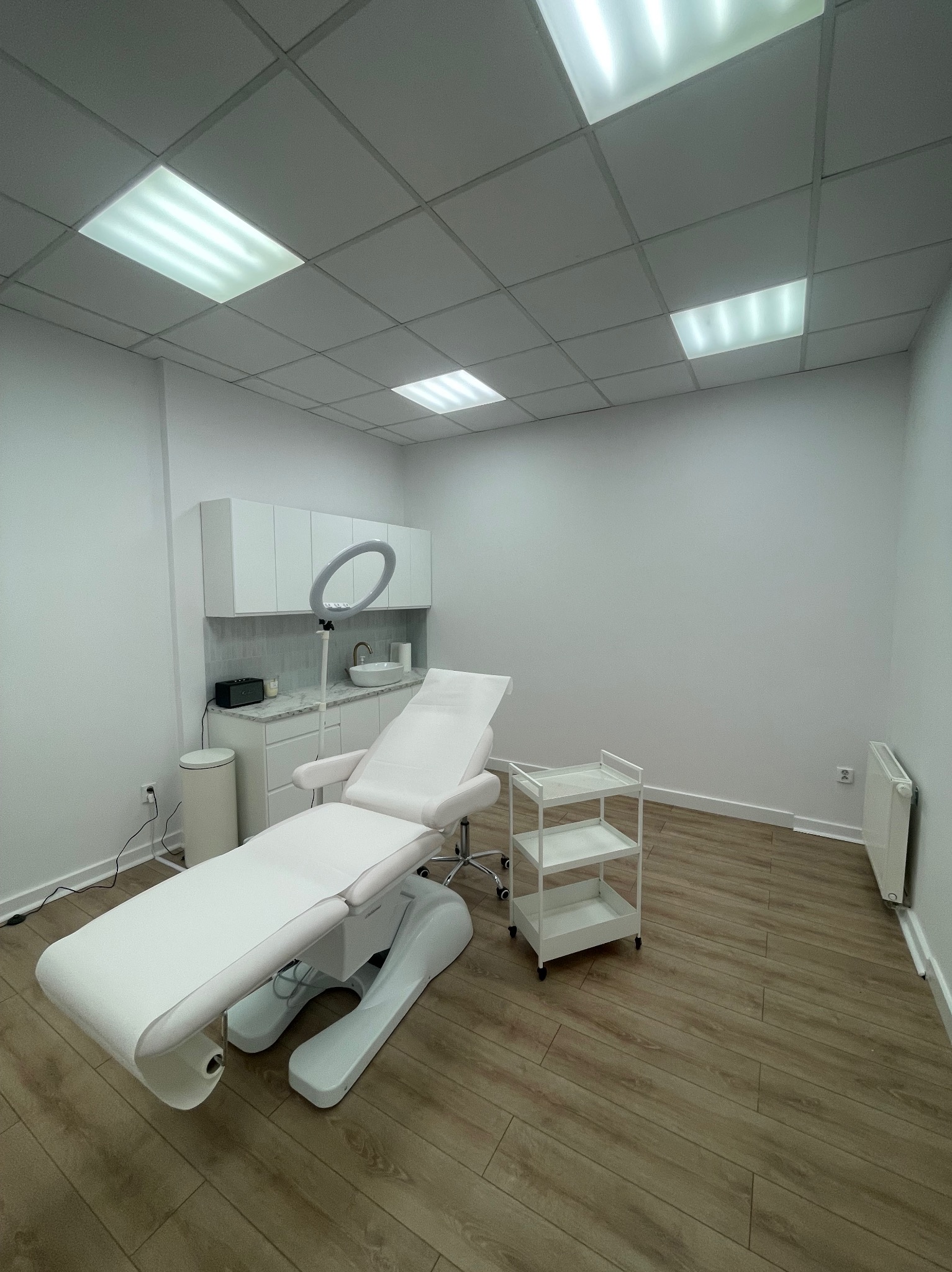 Jasne wnętrze gabinetu kosmetycznego z białym fotelem zabiegowym, szafkami, umywalką i oświetleniem LED. Podłoga z paneli imitujących drewno.