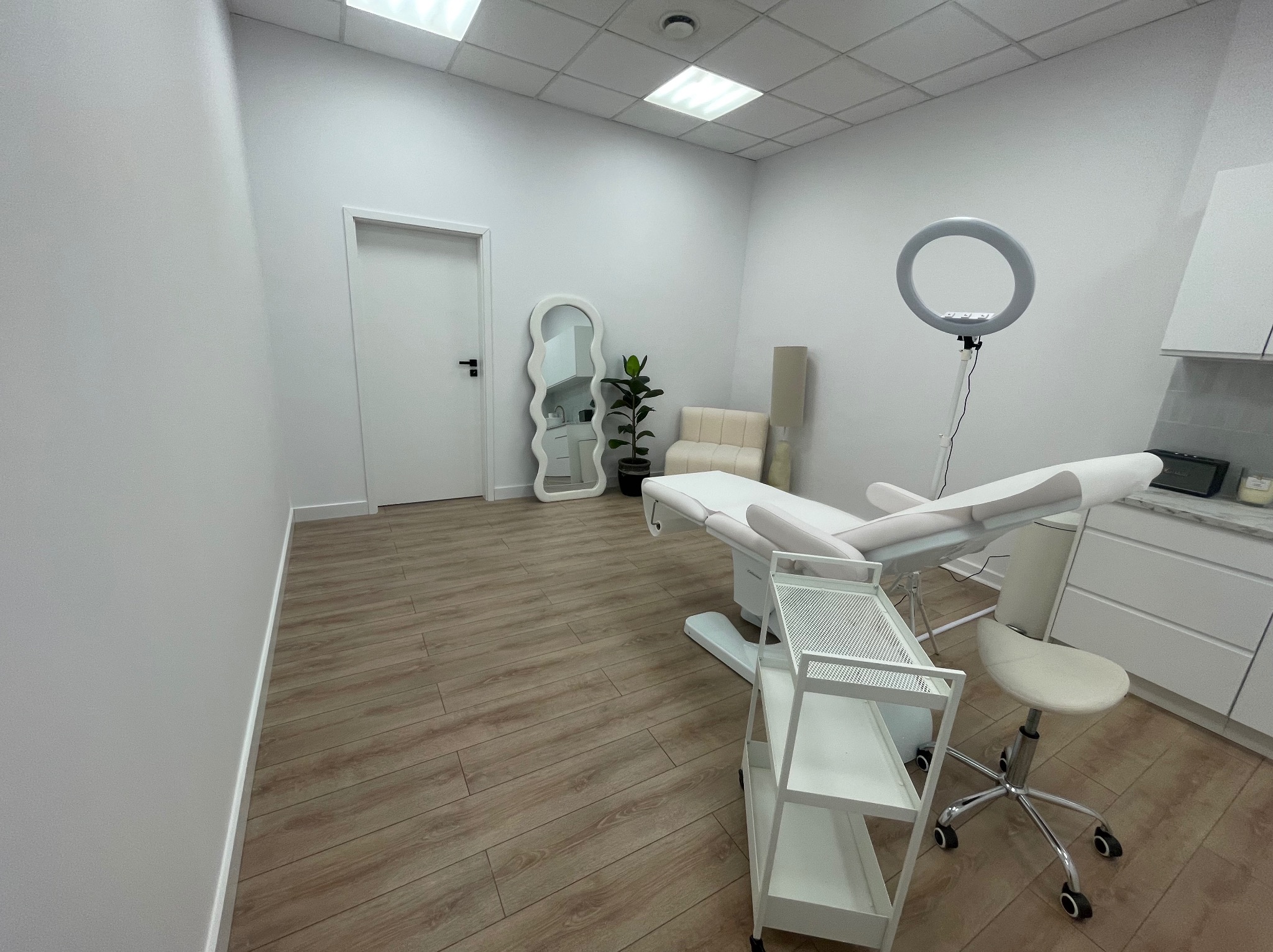 Jasne wnętrze gabinetu kosmetycznego z białym fotelem zabiegowym, lampą pierścieniową i drewnianą podłogą. Minimalistyczny design z lustrem i rośliną w tle.