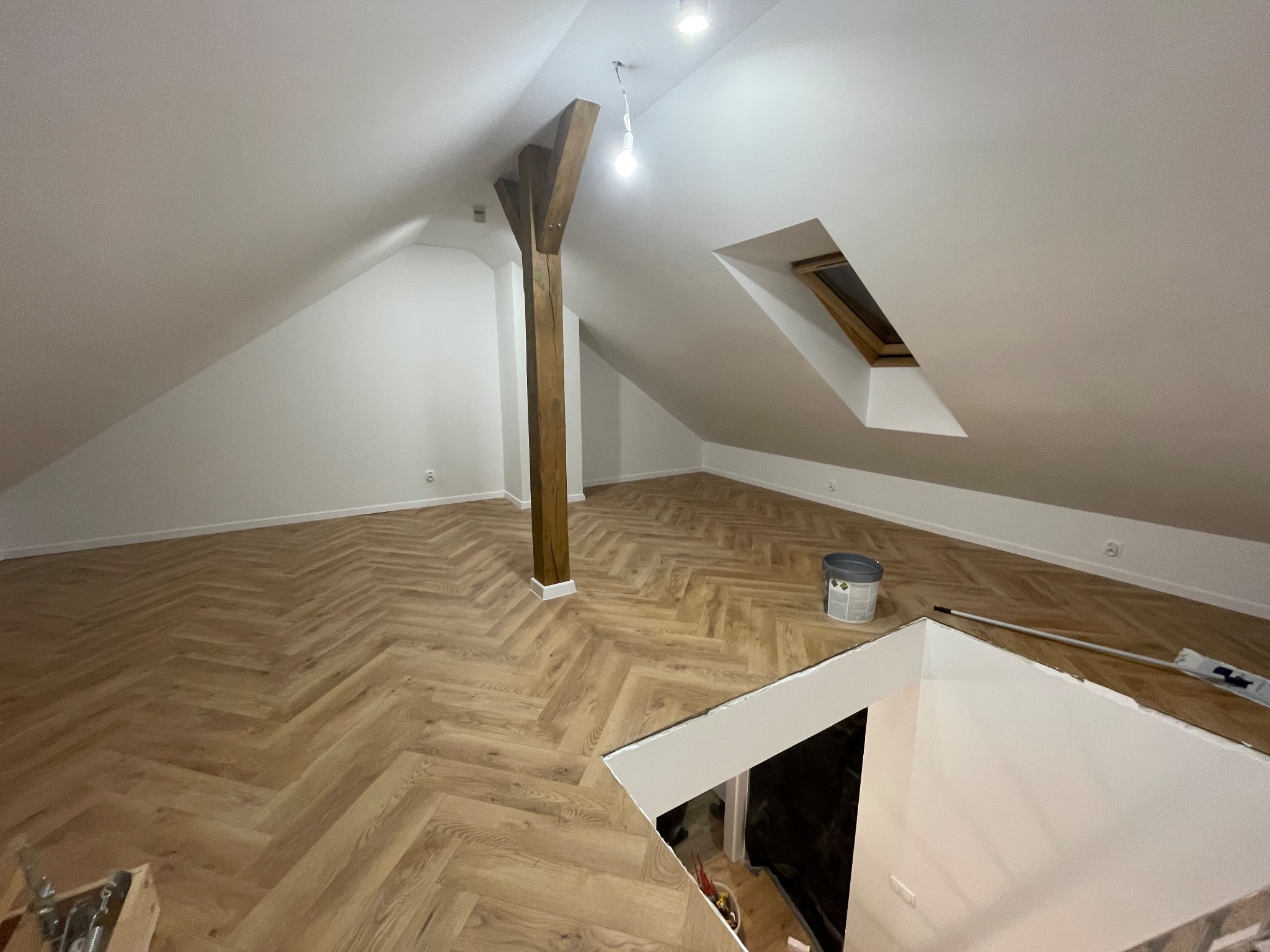 Poddasze po remoncie: parkiet w jodełkę, drewniany słup, okno dachowe. Widoczny fragment schodów i wiadro z farbą. Świeżo wykończone wnętrze.