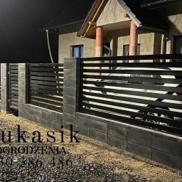 ŁUKASIK SPÓŁKA Z OGRANICZONĄ ODPOWIEDZIALNOŚCIĄ - Nowoczesne ogrodzenie z poziomych, czarnych paneli i szarych, betonowych słupków, oświetlone nocą, z widocznym logotypem firmy 'Łukasik Ogrodzenia' w lewym dolnym rogu.