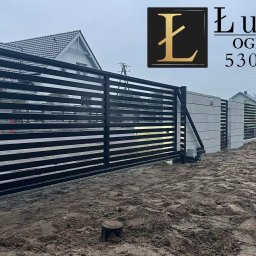ŁUKASIK SPÓŁKA Z OGRANICZONĄ ODPOWIEDZIALNOŚCIĄ - Automatyczna brama przesuwna z poziomych profili, szara podmurówka z logo firmy Łukasik Ogrodzenia i numerem telefonu, teren po montażu.