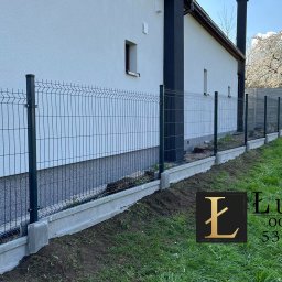 ŁUKASIK SPÓŁKA Z OGRANICZONĄ ODPOWIEDZIALNOŚCIĄ - Nowoczesne ogrodzenie panelowe w kolorze grafitowym, zamontowane na betonowej podmurówce, przylegające do białego budynku z ciemnymi elementami architektonicznymi, w tle kwitnące drzewo.