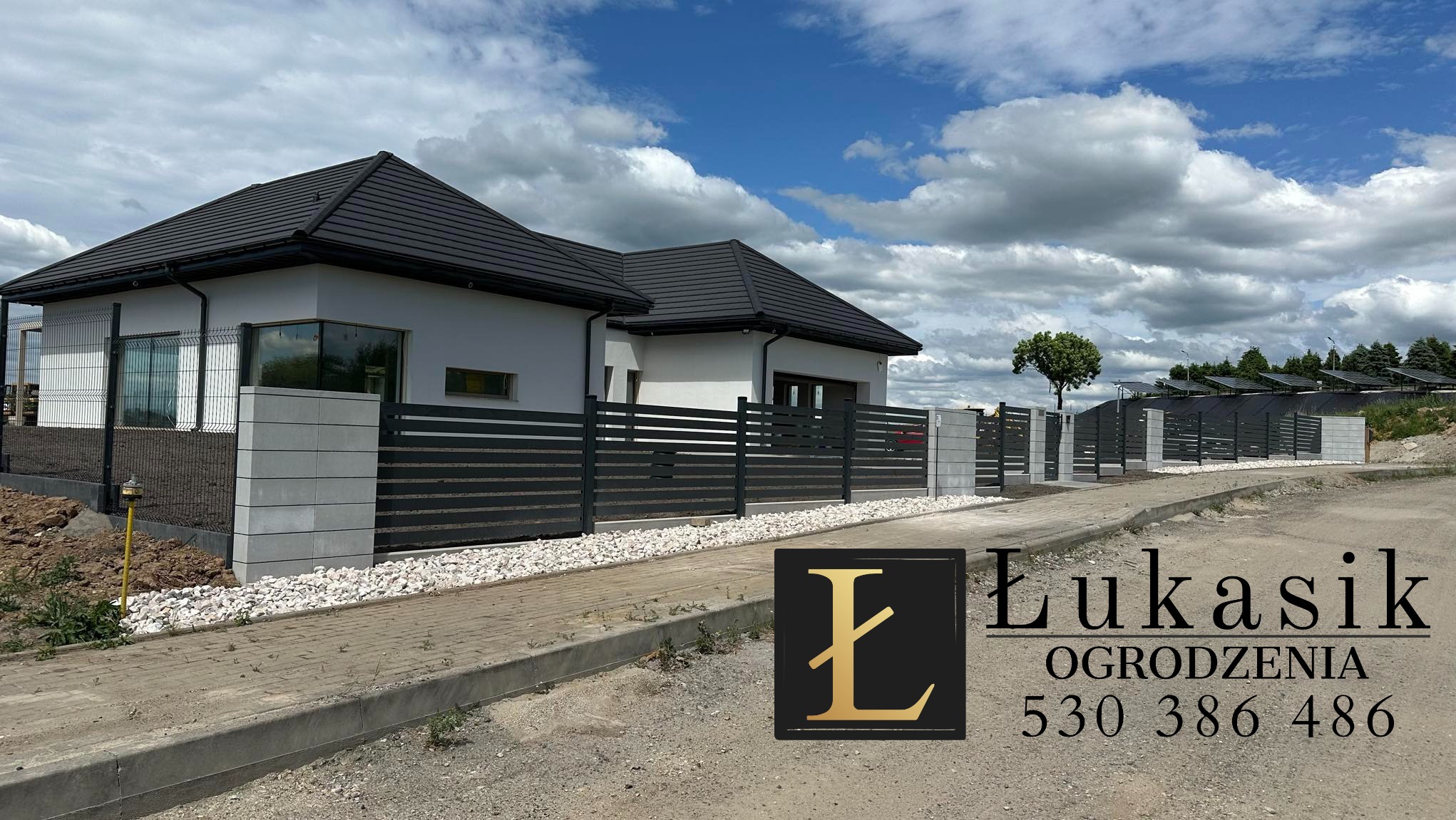 Nowoczesne ogrodzenie panelowe w kolorze antracytowym z betonowymi słupkami, wykończone białym kamieniem, na tle domu z ciemną dachówką i błękitnego nieba z chmurami.