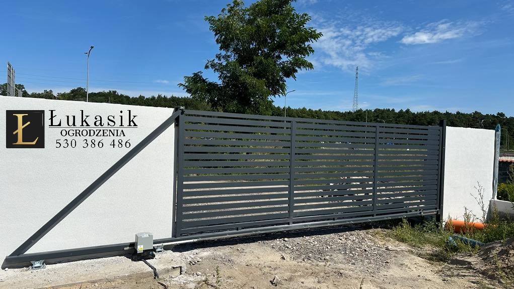Nowoczesna, przesuwna brama w kolorze grafitowym z automatyką, zamontowana przy białym, otynkowanym murze. Widoczne logo firmy Łukasik Ogrodzenia.