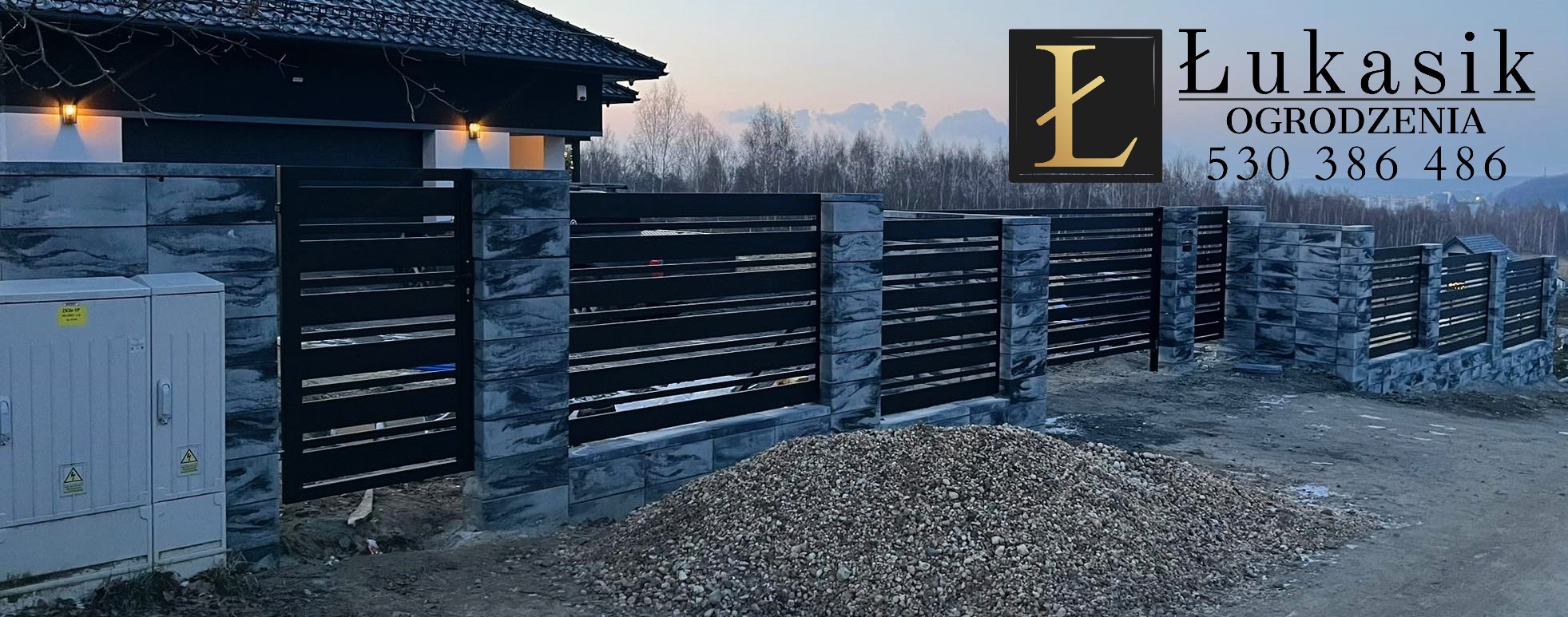 Nowoczesne ogrodzenie z poziomych, czarnych paneli między słupkami z szarego, ciosanego kamienia, widoczne logo firmy Łukasik Ogrodzenia, z przodu pryzma gruzu.
