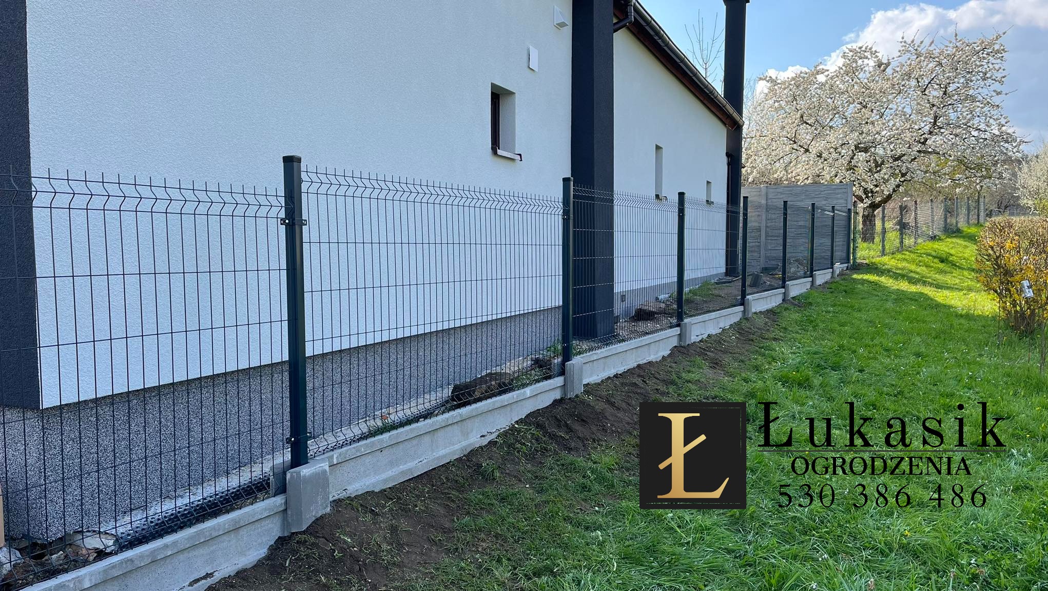 Nowoczesne ogrodzenie panelowe w kolorze grafitowym, zamontowane na betonowej podmurówce, przylegające do białego budynku z ciemnymi elementami architektonicznymi, w tle kwitnące drzewo.