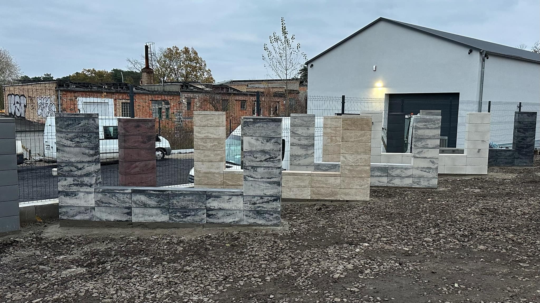 Wystawka fragmentów ogrodzenia z bloków betonowych o różnej kolorystyce i fakturze, na tle budynku gospodarczego i starej cegielni.