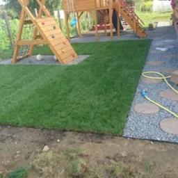 Świeżo ułożona, intensywnie zielona darń graniczy z placem zabaw dla dzieci i drewnianym tarasem z kamienną ścieżką, podlewane żółtym wężem ogrodowym.