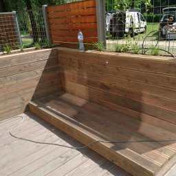 GREWE PAWEŁ CIURA - Drewniany taras z wbudowaną donicą i siedziskiem, widoczne narzędzia pracy i samochód dostawczy w tle, butelka wody na donicy.