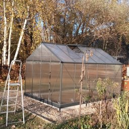 GREWE PAWEŁ CIURA - Przezroczysta szklarnia z otwartym oknem dachowym, ustawiona na żwirowym podłożu, z drabinką opartą o ścianę, na tle jesiennych drzew i budynku.