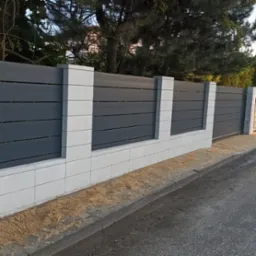 Nowoczesne ogrodzenie posesji: szare, poziome panele między białymi, kwadratowymi słupkami z bloczków. Widok na ulicę i drzewa w tle.