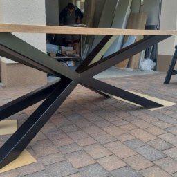 GREWE PAWEŁ CIURA - Stół z drewnianym blatem i metalową podstawą w kształcie gwiazdy, ustawiony na kostce brukowej. W tle widoczne narzędzia i materiały budowlane, co sugeruje montaż lub wykończenie.