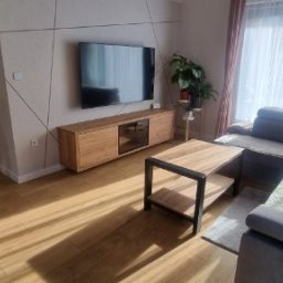 GREWE PAWEŁ CIURA - Jasne wnętrze salonu po remoncie: drewniana podłoga, szara sofa narożna, szafka RTV i TV na ścianie z dekoracyjnymi liniami. Widoczny odkurzacz w tle.