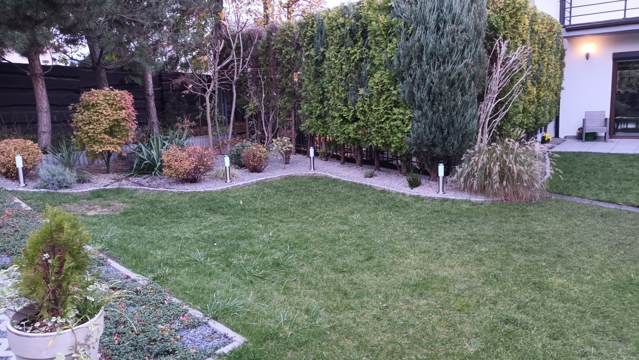 Zadbany ogród z równo przystrzyżonym trawnikiem, rabatami wysypanymi żwirem, oświetleniem ogrodowym i wysokimi tujami w tle, widok na taras i część domu.