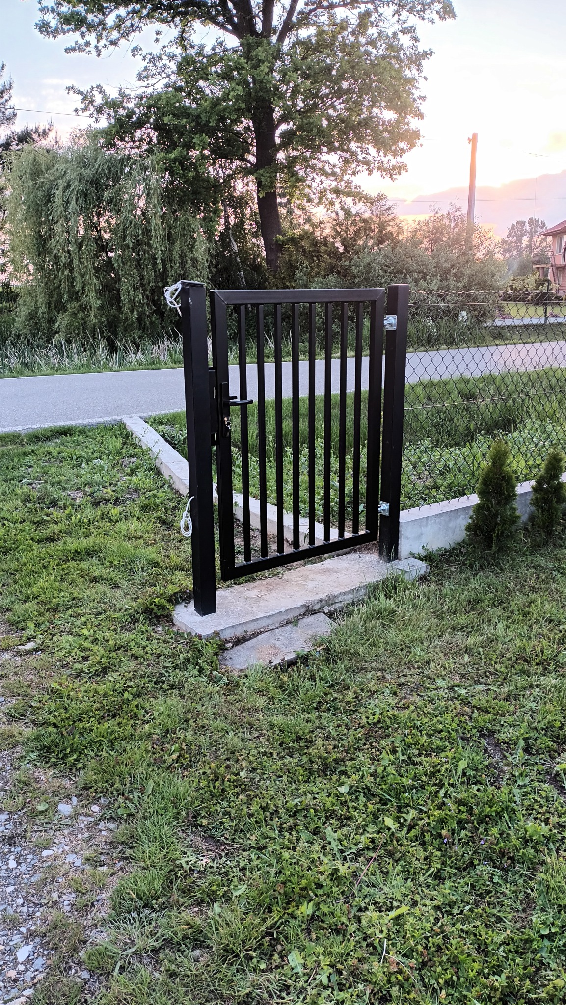 Czarna furtka ogrodowa z pionowymi szczebelkami, zamontowana na betonowej podstawie, otoczona zieloną trawą i ogrodzeniem z siatki.